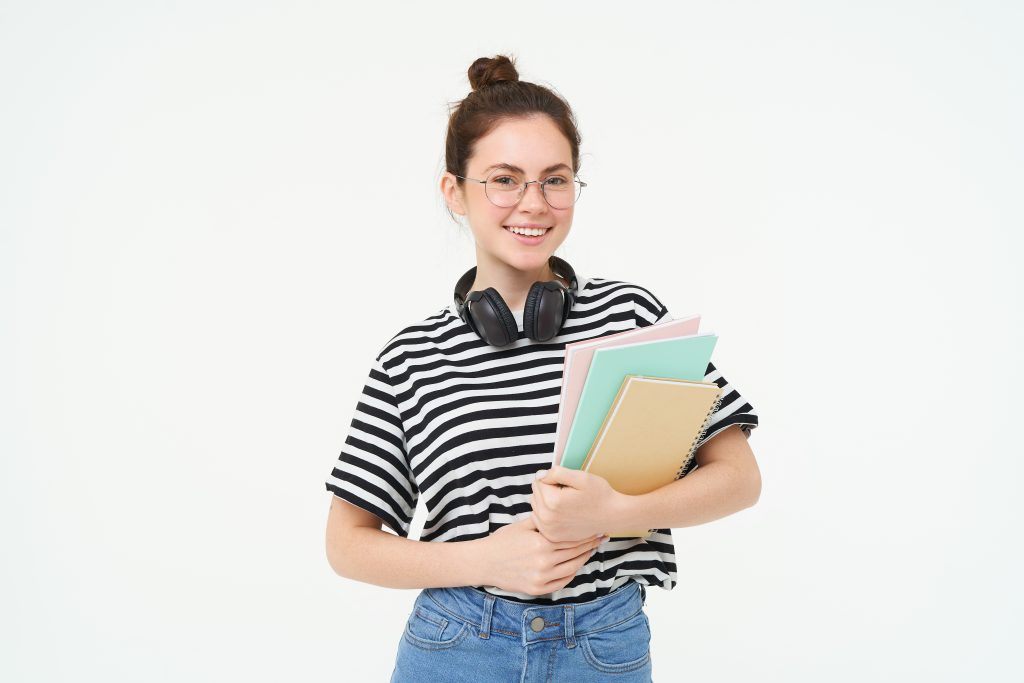 Education concept. Smiling brunette girl, student in casual clothes, holds her books, study material, wears headphones over neck, looks confident and happy, isolated over white background